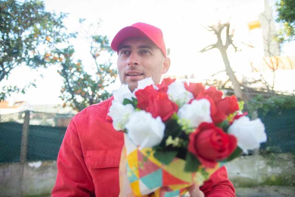 A young male delivery person wearing a bright yellow high-visibility vest and a red cap is outdoors on a residential street during daytime, carefully handling a fresh bouquet of white hydrangeas with