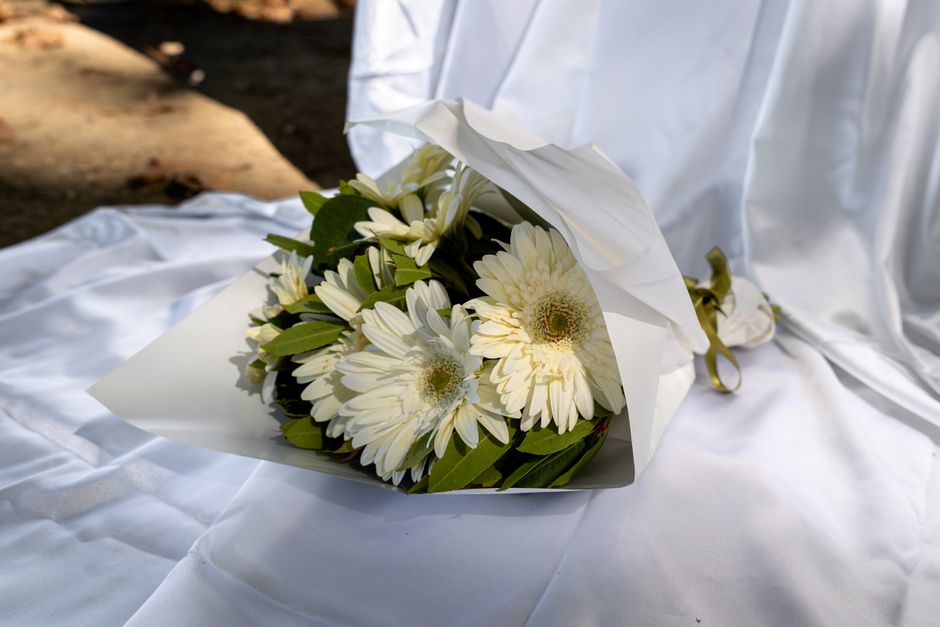 A bouquet of fresh white Gerbera daisies with dark centers, surrounded by green leaves, wrapped in white floral paper with a golden ribbon. The bouquet is placed on a white satin cloth, with natural l
