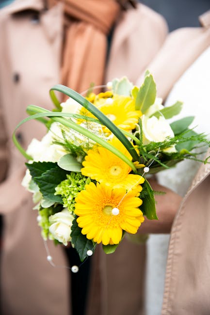 A florist from flowerstottenhamhale.co.uk is holding a fresh floral bouquet featuring vibrant yellow gerbera daisies, complemented by white roses and soft green foliage. The arrangement is natural and