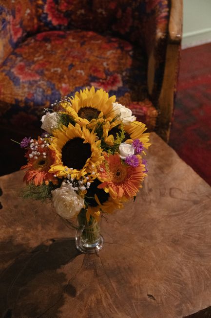 A vibrant floral bouquet featuring bright yellow sunflowers as the focal point, surrounded by smaller orange, pink, and purple flowers, including daisies and baby's breath, all arranged in a clear gla