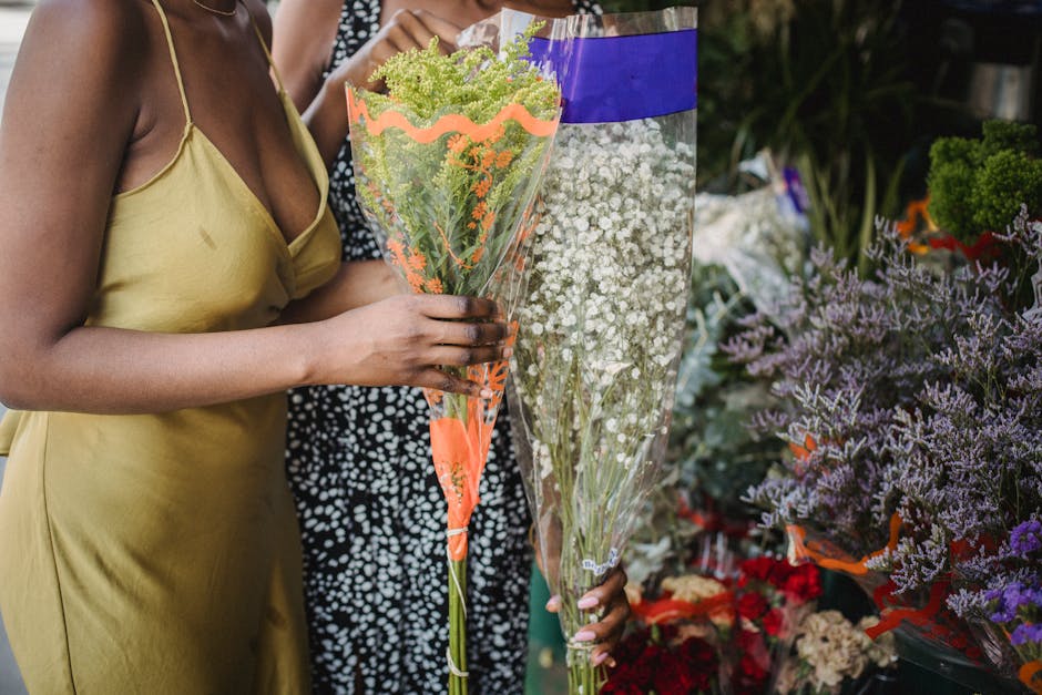 A woman in a yellow satin slip dress is holding a tall bouquet wrapped in clear and orange cellophane, featuring green and purple flowers, possibly greenery and small blossoms. She is standing in a fl