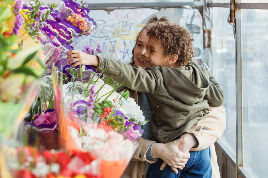 A young boy with curly hair, wearing a green hoodie, is smiling and reaching out to pick a colorful floral bouquet at a flower shop. The arrangement includes vibrant pink, purple, white, and yellow fl